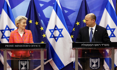 Ursula von der Leyen smiles at Naftali Bennet while they stand at podiums with EU and Israel flags behind them