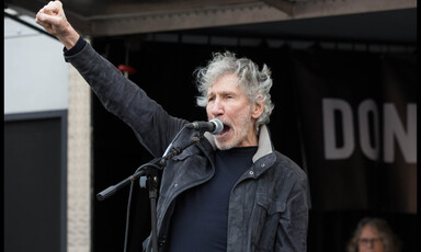 Roger Waters, dressed in a black t-shirt and a black jacket, stands at a microphone with his fist in the air