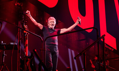 Roger Waters, dressed in a black t-shirt and black jeans and holding a microphone, raises his arms and smiles onstage