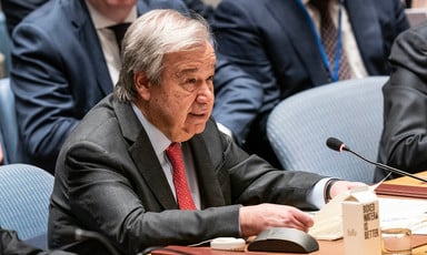 António Guterres holds a piece of paper while sitting at conference table in front of microphone