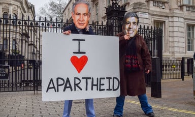 Two people wearing masks mimicking Benjamin Netanyahu and Rishi Sunak hold a sign reading I heart apartheid 