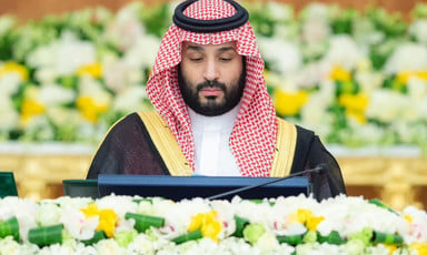 Bin Salman, wearing a head dress and a gold-trimmed cloak, looks down towards a podium while surrounded by flowers