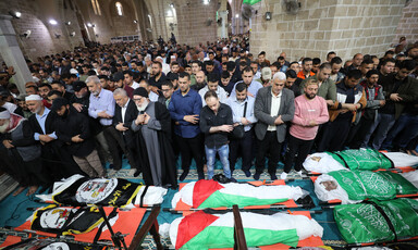 Men stand while praying while facing bodies wrapped in Palestine and faction flags on ground in front of them inside mosque