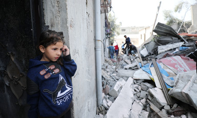 Child wearing a navy sweatshirt stands in the doorway of a building beside a pile of rubble 