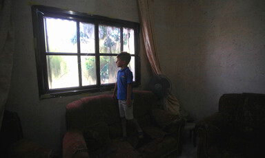 A small boy in dark room looks out of window with broken glass