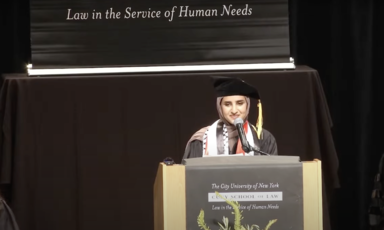 Fatima Mohammed, dressed in a graduation cap and gown, and wearing traditional Palestinian scarves,  stands at a podium.