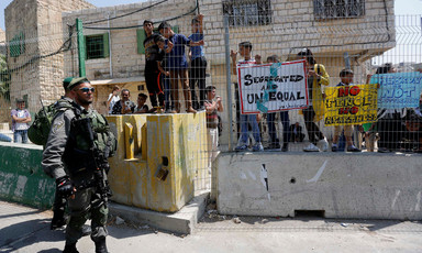 Israeli soldiers stand in front of a group of children who are behind a fence on a street. They are holding signs that denounce apartheid and segregation.