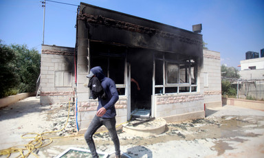 Man holds cloth to his face while walking in front of scorched single-story residential building with its front door and windows destroyed
