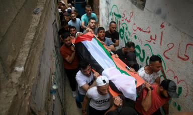 Men in a narrow alleyway carry a stretcher draped in a Palestinian flag