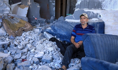 Man wearing a baseball cap sits amid the rubble of a destroyed building 