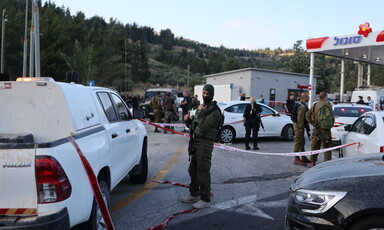 Soldier wearing gaiter over his face looks toward camera while standing in front of area marked with ribbon at gas station as other armed personnel stand around