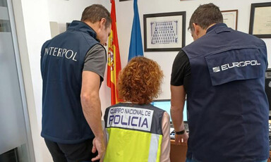 Two people, one wearing a vest reading Interpol, the other wearing a vest reading Europol stand on either side of a seated person wearing a vest reading Cuerpo Nacional de Policia