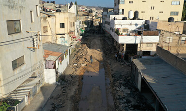 Man seen from above walks along a wide dug-up road with residential buildings on opposite sides