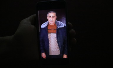 A cell phone photograph depicting Amin Warda in a jacket surrounded by a dark background.