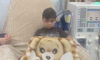 A boy wrapped in a blanket depicting a large teddy bear sits on a chair beside a dialysis machine