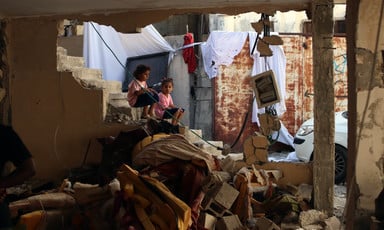 Two young girls sit on stairs in debris-strewn home with a wall blown open