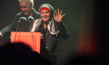 The singer Sinéad O'Connor stands in front of a microphone and waves 