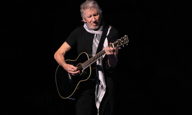 The musician Roger Waters wears a black and white checkered scarf while playing guitar onstage