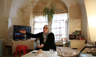 Woman sitting in front of table with arched window behind her raises her right hand while talking