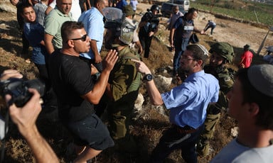 Man wearing kippah and civilian clothing points at another civilian with his hand in front of his face as soldiers and others stand nearby