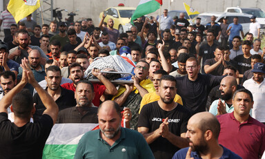Body shrouded in kuffiyeh and Palestine flag is carried on stretcher by crowd of men, some carrying Palestine and Fatah flags