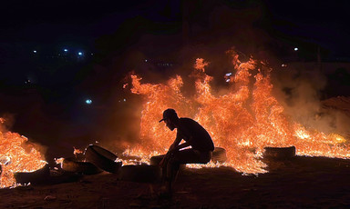 The silhouette of a man wearing a cap against the backdrop of fire and burning tires 