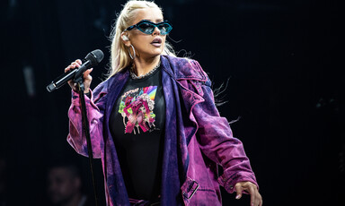 Christina Aguilera, in a purple jacket and wearing sunglasses, holds a microphone