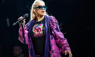 Christina Aguilera, in a purple jacket and wearing sunglasses, holds a microphone