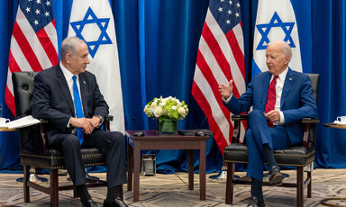 Joe Biden raises his right hand while sitting in chair opposite from a seated Netanyahu with US and Israeli flags behind them