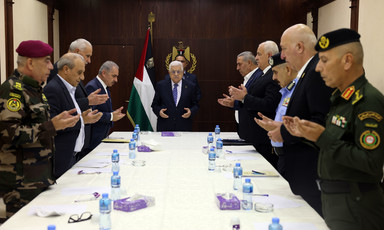 Men, some in uniform and others in suits, stand around a meeting table with Abbas at its head