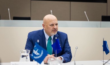 The International Criminal Court's chief prosecutor Karim Khan sits at a desk with microphones, blue and white flags and two jugs of water  desk with 