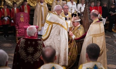 Welby in gold-trimmed bishop's robes places ornate crown on head of seated, robed monarch Charles as others look on
