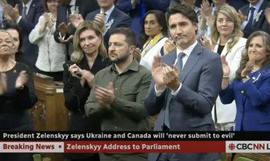 Zelensky in a military shirt stands next to Trudeau who is wearing a suit and clapping as others look on