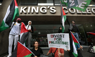 People holding signs and flags appear to be listening to a speaker who is not visible in the picture