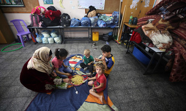 A woman and four children sit on rugs on a floor 