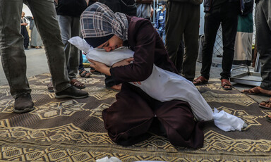 A young woman cries while holding her arms tight around a shrouded body and kneeling on the ground