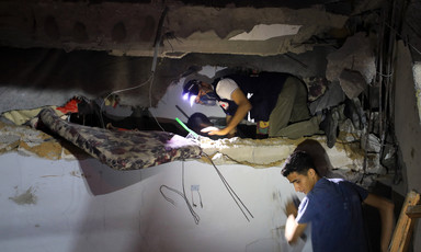 A man wearing a helmet with a light on it searches a building that has been bombed in Gaza City, accompanied by another person who is not wearning protective headgear  