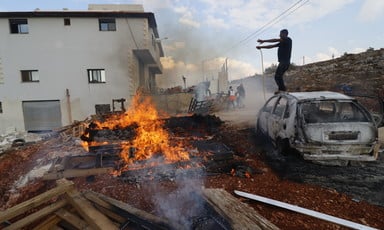 A fire blazes beside a burnt car amidst the destruction caused by a nearby building.