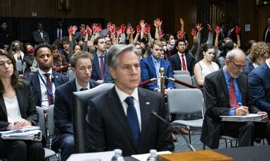 Man in a suit is seated while audience behind him has their hands raised and painted red 