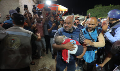 Wael Dahdouh carries a toddler while wearing a flak jacket and as other journalists stand around him, many with cameras