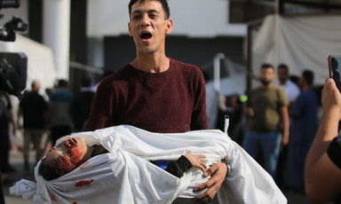 Young man with his mouth open carries the body of a child wrapped loosely in a white sheet with a bloodied head and closed eyes