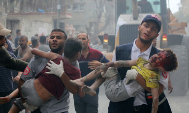 Two men carry children covered in dust and blood amid ruined buildings and chaos