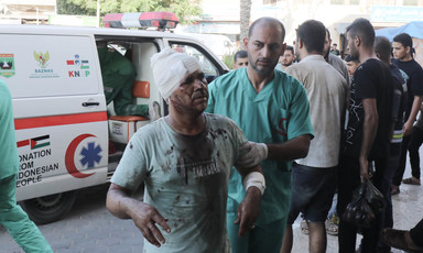 A wounded man with his head wrapped in a bandages is brought into hospital by a medic wearing green overalls