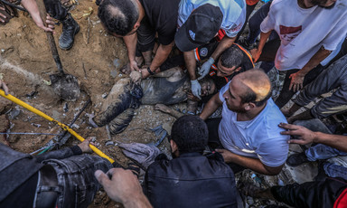 Men, most with bare hands, hold the body of a man partially buried in sandy rubble