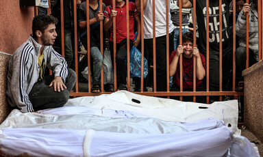 A man cries as he sits next to several shrouded bodies with mourning people standing on opposite side of metal fence behind him