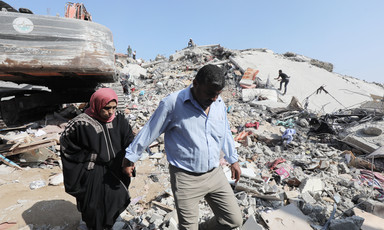 A woman wearing a red head scarf and black clothes and a man wearing a blue shirt and beige trousers walk amid the remains of a building that has been destroyed 