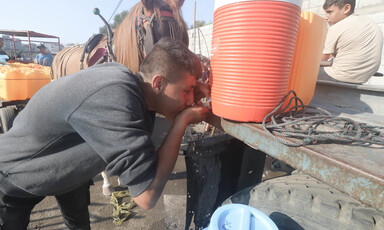 A boy positions himself under an orange water jug to drink water from its spout.