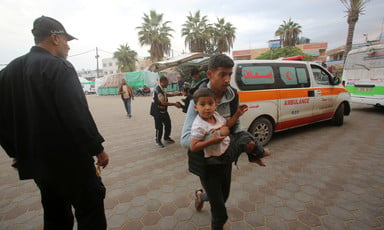 A medic carries a boy toward a hospital in Gaza. An ambulance can be seen behind the medic