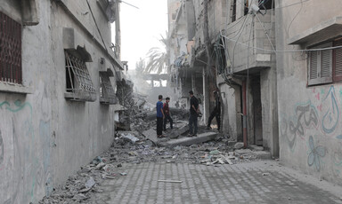 Four men stand along a small road between destroyed homes covered in dust