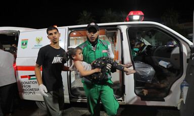 A medic wearing a baseball cap and green overalls carries a girl in front of an ambulance 